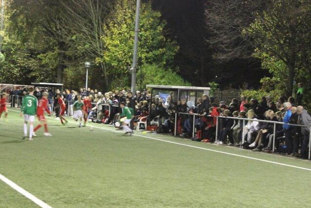 Spielszene auf einem beleuchteten Fu&szlig;ballplatz mit Spielern in gr&uuml;nen und roten Trikots sowie zahlreichen Zuschauern am Rand.