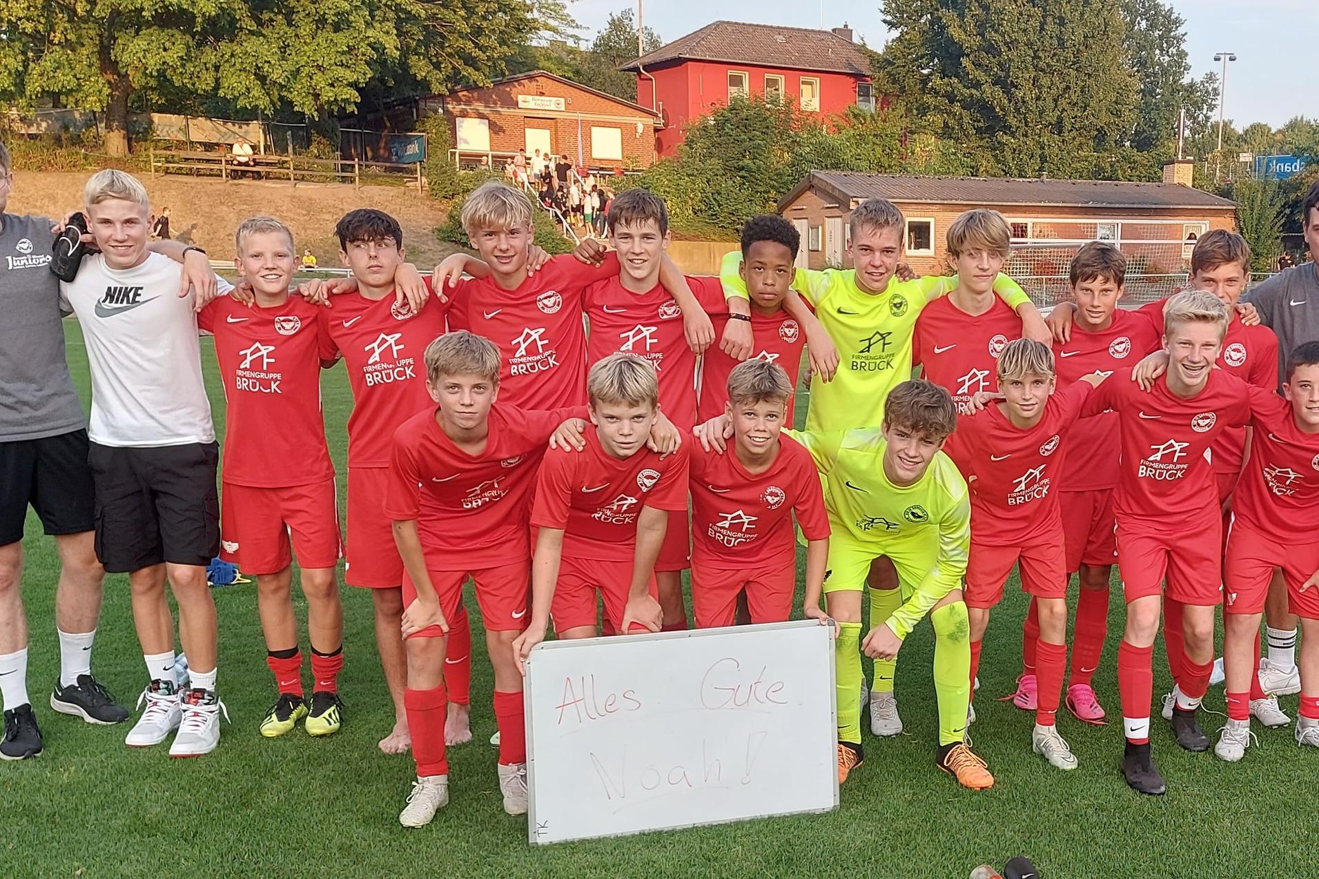 Jugendfu&szlig;ballmannschaft in roten Trikots posiert mit Trainern auf einem Platz, daneben ein Schild mit Schriftzug.
