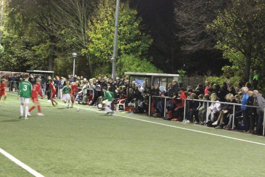 Spielszene auf einem beleuchteten Fu&szlig;ballplatz mit Spielern in gr&uuml;nen und roten Trikots sowie zahlreichen Zuschauern am Rand.