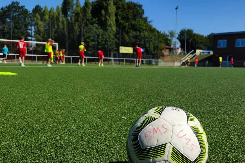 Nahaufnahme eines Fu&szlig;balls auf einem Kunstrasenplatz mit Spielern im Hintergrund, die ein Training absolvieren.