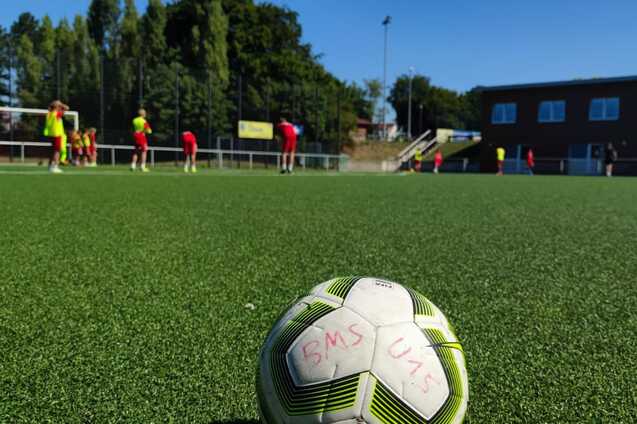 Nahaufnahme eines Fu&szlig;balls auf einem Kunstrasenplatz mit Spielern im Hintergrund, die ein Training absolvieren.