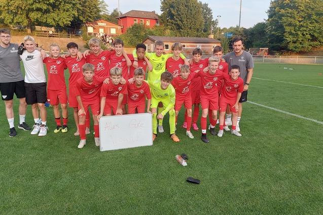 Jugendfu&szlig;ballmannschaft in roten Trikots posiert mit Trainern auf einem Platz, daneben ein Schild mit Schriftzug.