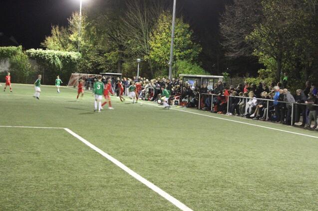 Spielszene auf einem beleuchteten Fu&szlig;ballplatz mit Spielern in gr&uuml;nen und roten Trikots sowie zahlreichen Zuschauern am Rand.