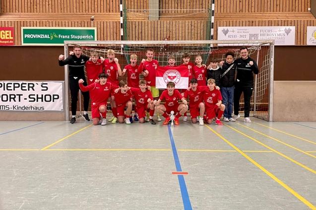 Gruppe von Jugendlichen in roten Trikots posiert jubelnd vor einem Handballtor in einer Sporthalle.