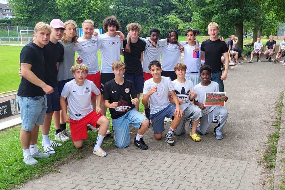 Gruppe von Jugendlichen in Sportkleidung posiert l&auml;chelnd auf einem Weg neben einem Fu&szlig;ballplatz.