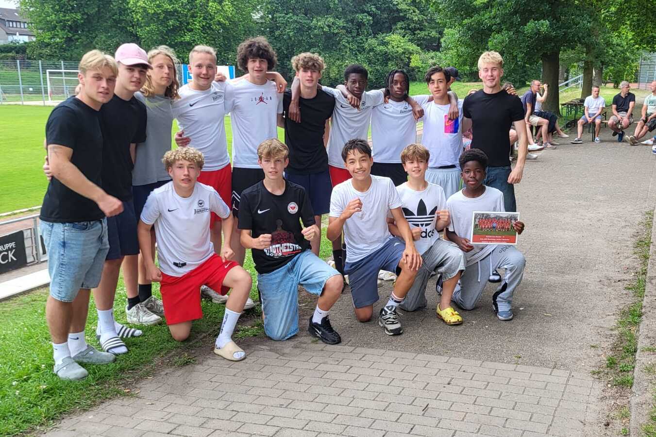 Gruppe von Jugendlichen in Sportkleidung posiert l&auml;chelnd auf einem Weg neben einem Fu&szlig;ballplatz.