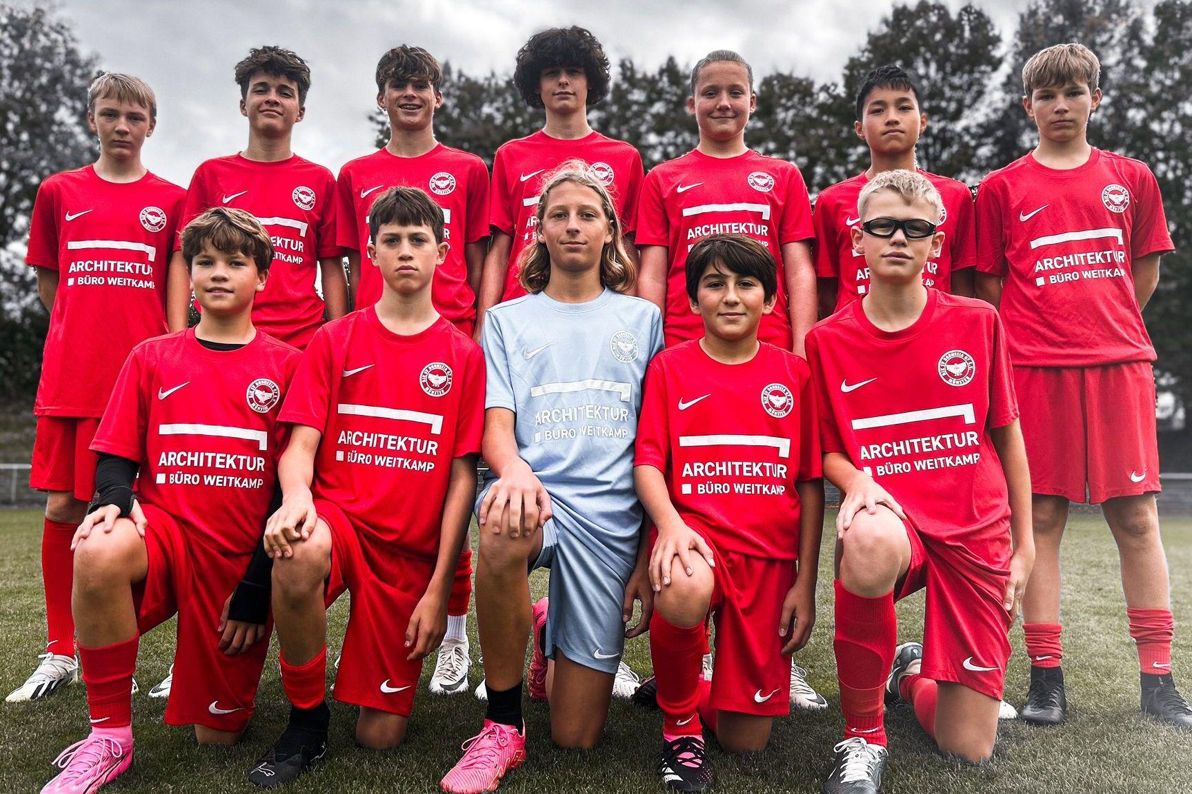 Elf Jugendfu&szlig;ballspieler in roten Trikots posieren auf einem Sportfeld, umgeben von B&auml;umen und einem bew&ouml;lkten Himmel.