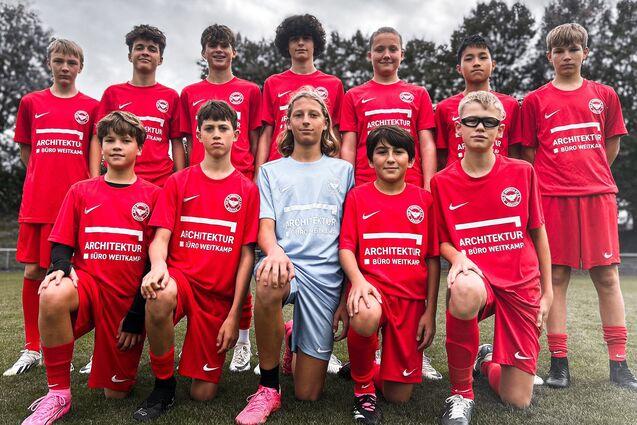 Elf Jugendfu&szlig;ballspieler in roten Trikots posieren auf einem Sportfeld, umgeben von B&auml;umen und einem bew&ouml;lkten Himmel.