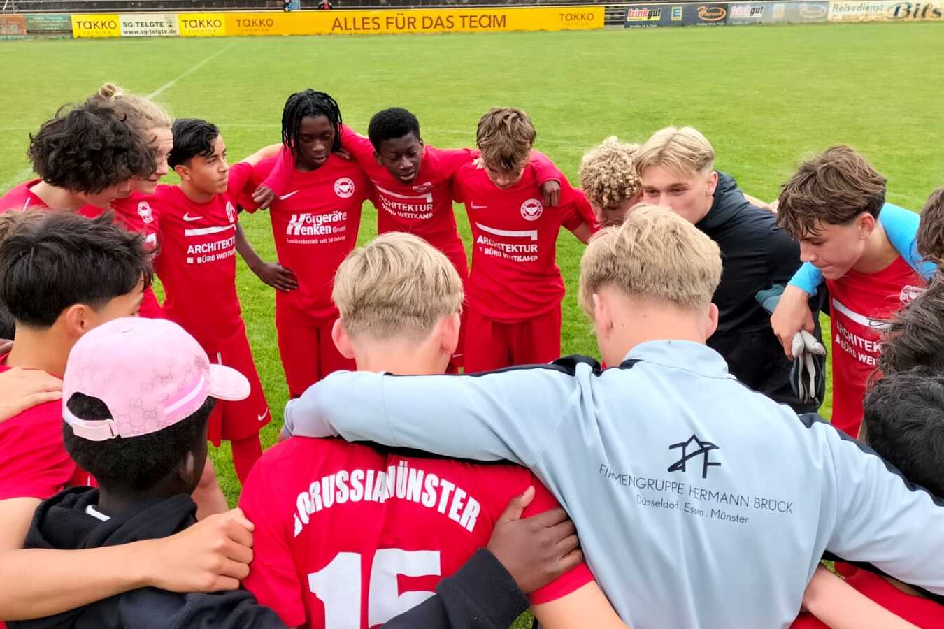 Gruppe von jugendlichen Fu&szlig;ballspielern in roten Trikots umringen einen Trainer auf dem Spielfeld.
