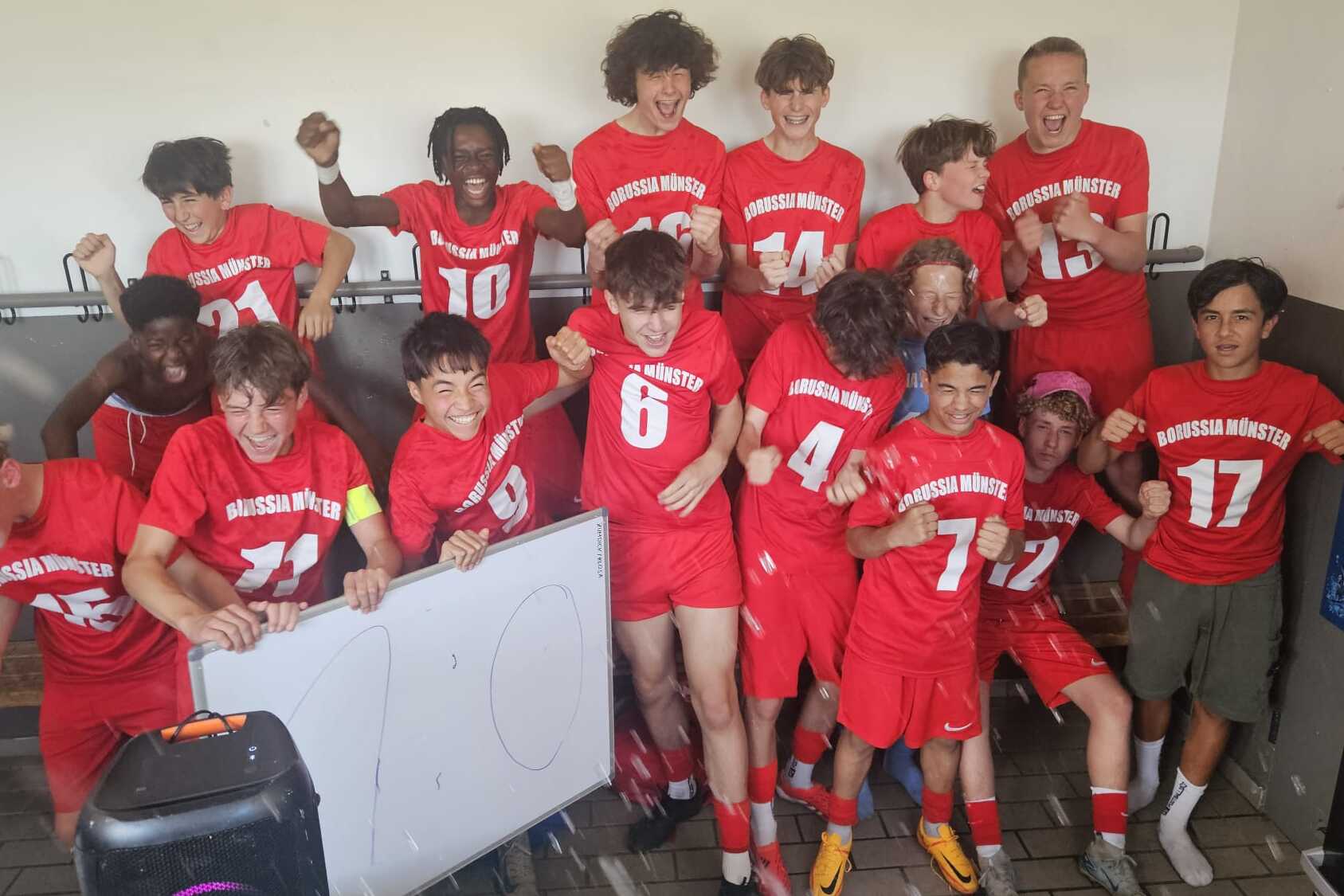 Gruppe junger Fu&szlig;ballspieler in roten Trikots posiert l&auml;chelnd und feiernd in einer Umkleidekabine.