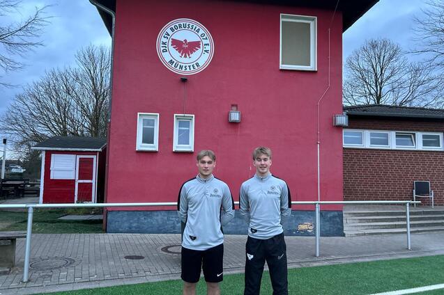 Zwei Jugendliche in Sportkleidung stehen vor einem roten Vereinsgeb&auml;ude mit dem Logo von Borussia M&uuml;nster.