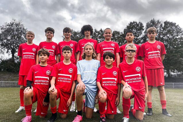 Elf Jugendfu&szlig;ballspieler in roten Trikots posieren auf einem Sportfeld, umgeben von B&auml;umen und einem bew&ouml;lkten Himmel.