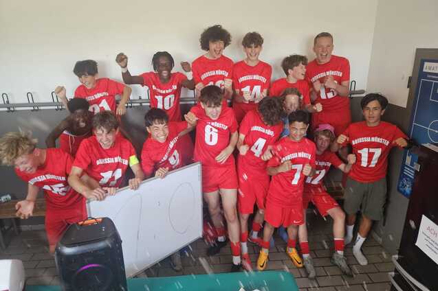 Gruppe junger Fu&szlig;ballspieler in roten Trikots posiert l&auml;chelnd und feiernd in einer Umkleidekabine.
