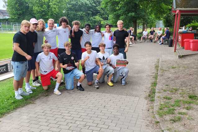 Gruppe von Jugendlichen in Sportkleidung posiert l&auml;chelnd auf einem Weg neben einem Fu&szlig;ballplatz.