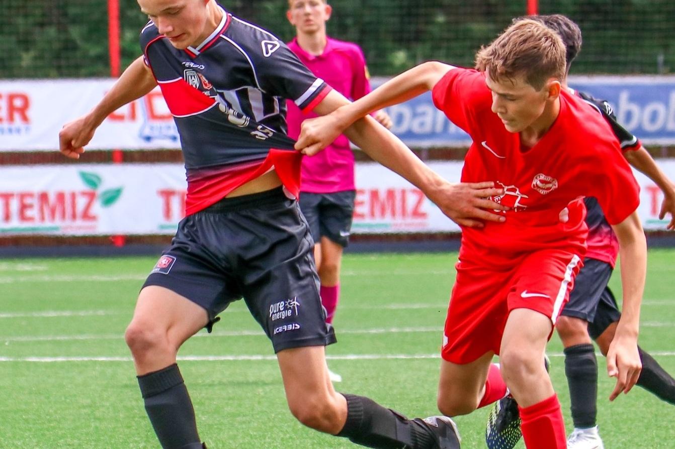 Zwei Jugendliche spielen Fu&szlig;ball auf einem Kunstrasenplatz, der Spieler in schwarz dribbelt den Ball, w&auml;hrend der in rot ihn versucht zu stoppen.