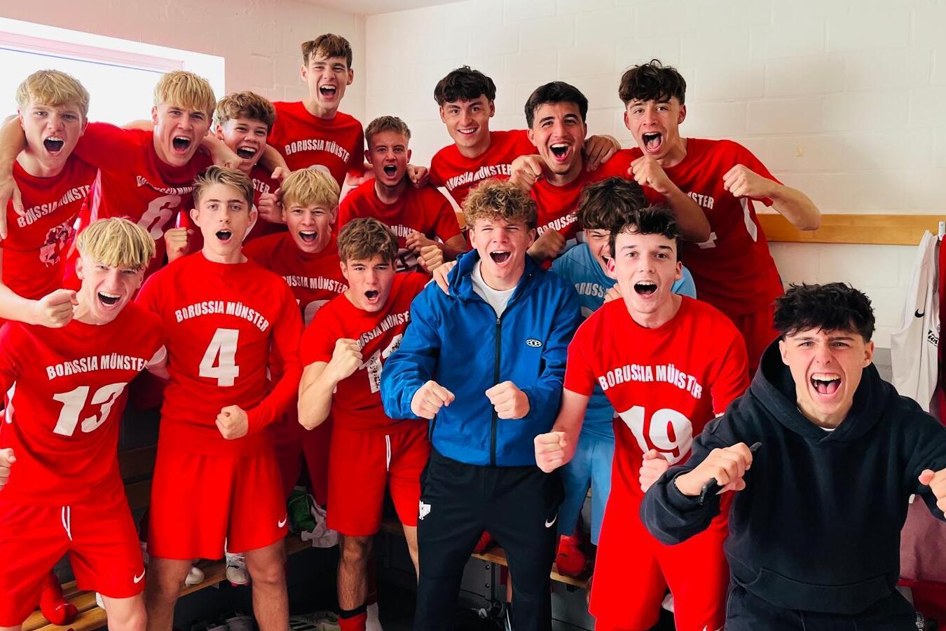 Gruppe von jubelnden jungen Fu&szlig;ballspielern in roten Trikots, die in einer Umkleidekabine stehen.