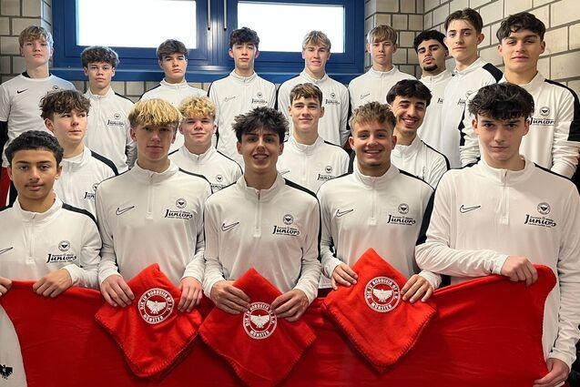 Gruppe von 16 jungen Fu&szlig;ballspielern in wei&szlig;en Trikots mit roten Flaggen, aufgereiht in einem Umkleideraum.
