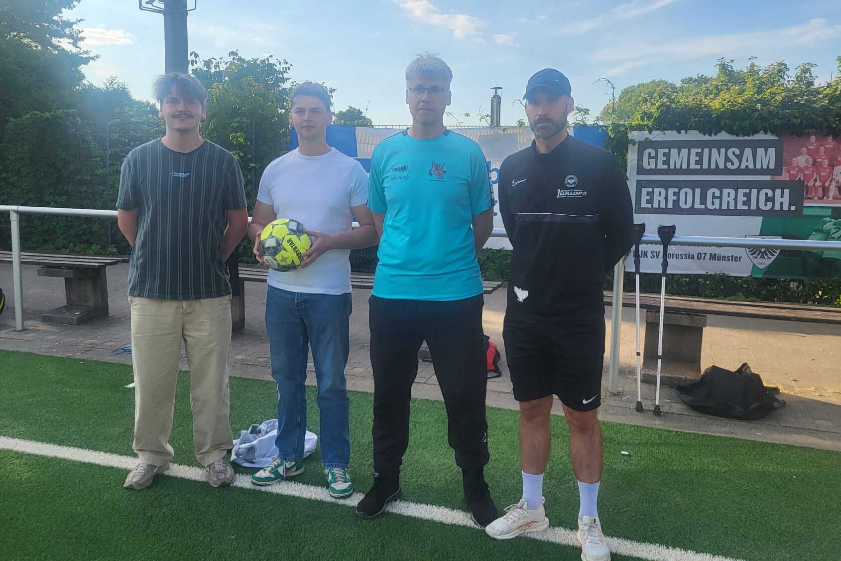 Vier M&auml;nner stehen auf einem Fu&szlig;ballplatz, einer h&auml;lt einen Ball, im Hintergrund ein Banner mit &bdquo;Gemeinsam erfolgreich&ldquo;.