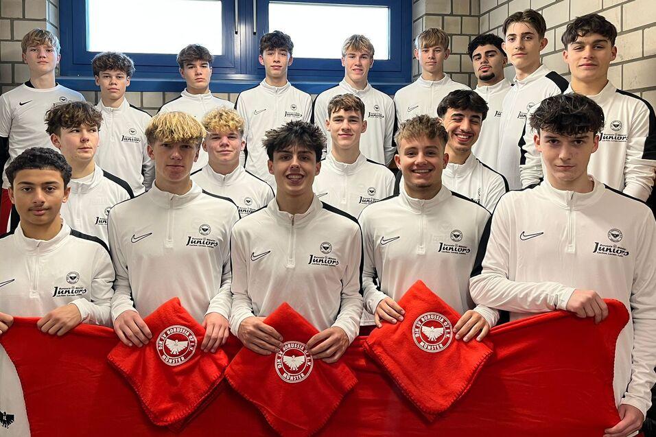 Gruppe von 16 jungen Fu&szlig;ballspielern in wei&szlig;en Trikots mit roten Flaggen, aufgereiht in einem Umkleideraum.