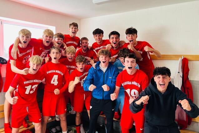 Gruppe von jubelnden jungen Fu&szlig;ballspielern in roten Trikots, die in einer Umkleidekabine stehen.
