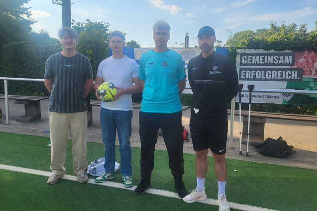 Vier M&auml;nner stehen auf einem Fu&szlig;ballplatz, einer h&auml;lt einen Ball, im Hintergrund ein Banner mit &bdquo;Gemeinsam erfolgreich&ldquo;.