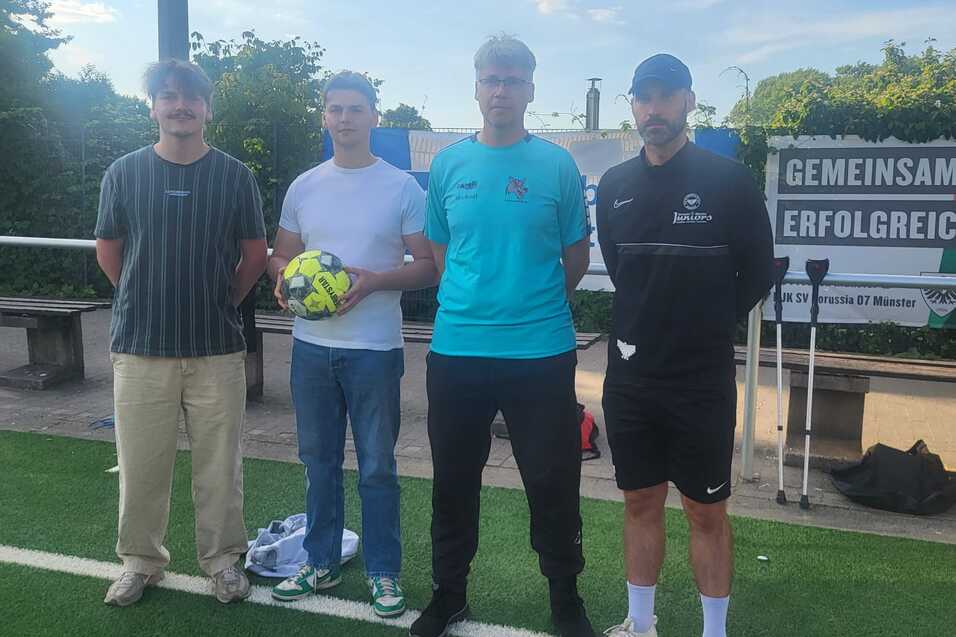 Vier M&auml;nner stehen auf einem Fu&szlig;ballplatz, einer h&auml;lt einen Ball, im Hintergrund ein Banner mit &bdquo;Gemeinsam erfolgreich&ldquo;.