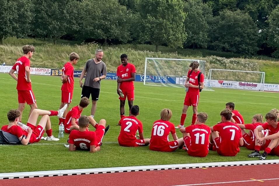 Eine Fu&szlig;ballmannschaft in roten Trikots sitzt und steht auf einer Wiese, umgeben von B&auml;umen und einem Sportplatz.