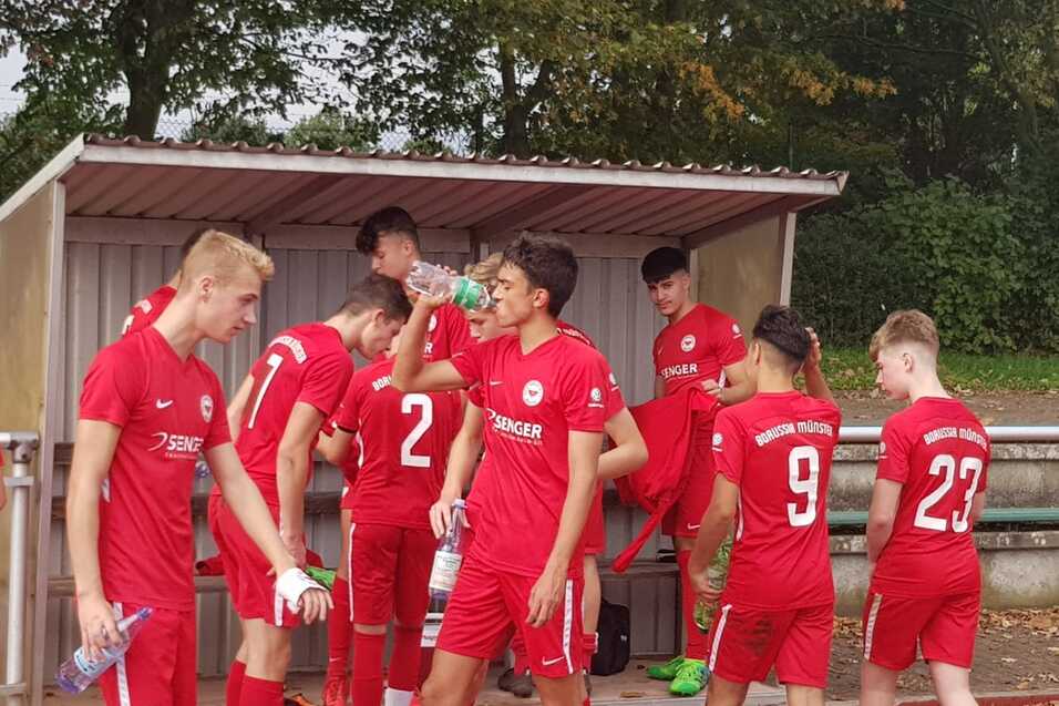 Gruppe junger Fu&szlig;ballspieler in roten Trikots versammelt sich in einer Kabine, einige trinken Wasser.