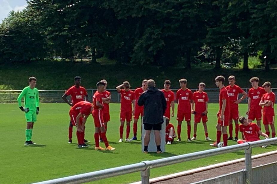 Eine Gruppe von Fu&szlig;ballspielern in roten Trikots versammelt sich um einen Trainer auf einem gr&uuml;nen Sportplatz.