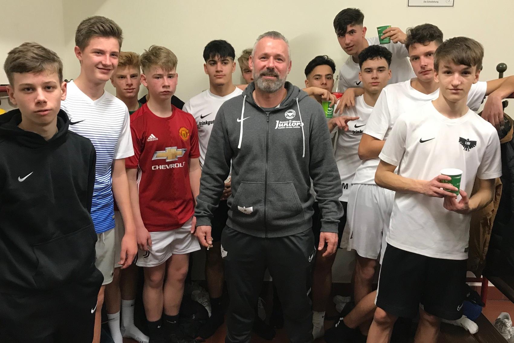 Gruppe von jungen Fu&szlig;ballspielern in einem Umkleideraum, umrahmt von einem Trainer in sportlicher Bekleidung.