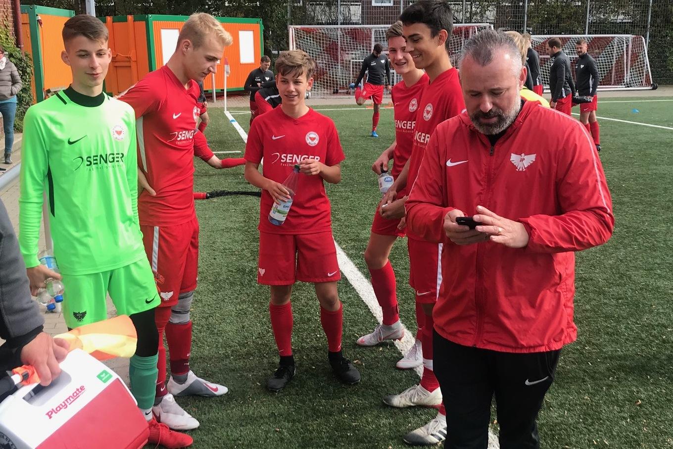 Gruppe von Jugendlichen in roten Fu&szlig;balltrikots auf dem Platz, Trainer bespricht sich mit ihnen.