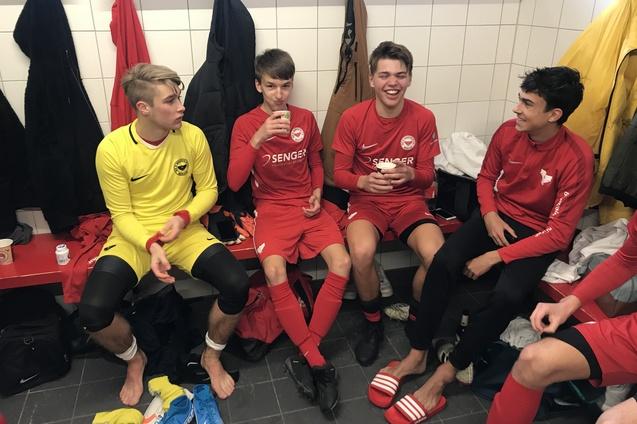 F&uuml;nf junge Fu&szlig;ballspieler in roter Sportkleidung sitzen in einer Umkleidekabine und unterhalten sich.