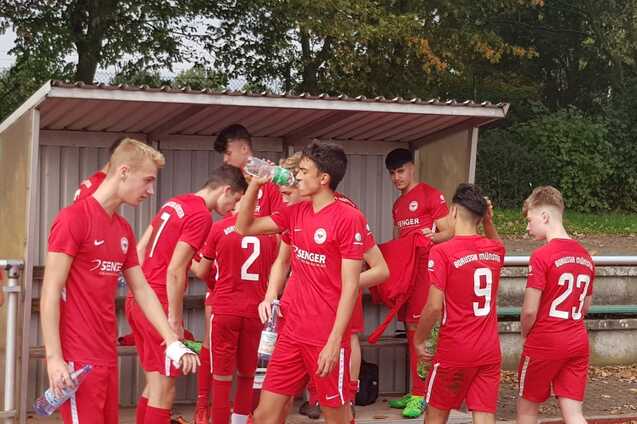 Gruppe junger Fu&szlig;ballspieler in roten Trikots versammelt sich in einer Kabine, einige trinken Wasser.