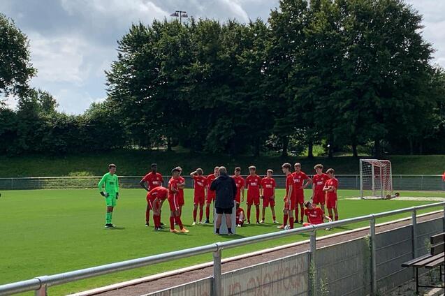 Eine Gruppe von Fu&szlig;ballspielern in roten Trikots versammelt sich um einen Trainer auf einem gr&uuml;nen Sportplatz.