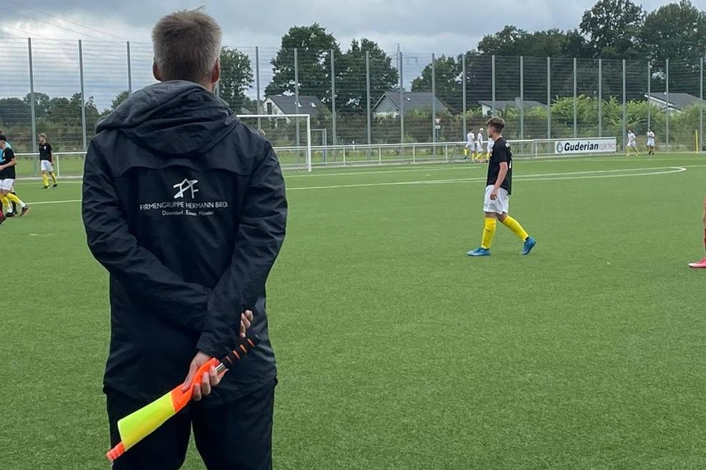 Ein Schiedsrichter mit Linienrichterflagge beobachtet ein Fu&szlig;ballspiel auf einem Kunstrasenplatz.