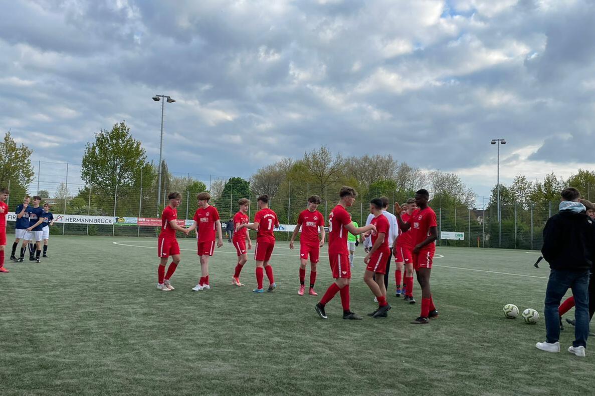 Fu&szlig;ballspieler in roten Trikots versammeln sich auf einem Kunstrasenplatz, im Hintergrund B&auml;ume und bew&ouml;lkter Himmel.