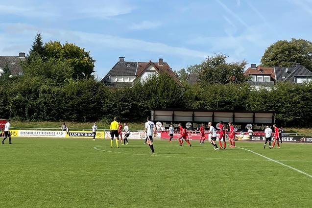 Fu&szlig;ballspiel auf gr&uuml;ner Wiese mit Spielern in roten und wei&szlig;en Trikots, im Hintergrund Wohnh&auml;user und B&auml;ume.