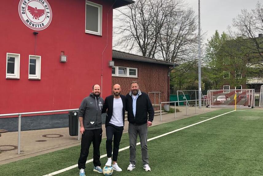 Drei M&auml;nner stehen auf einem Fu&szlig;ballplatz vor einem roten Vereinsgeb&auml;ude, mit einem Ball auf dem Rasen.