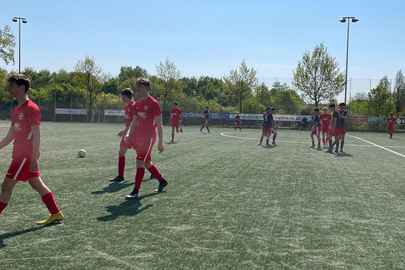 Fu&szlig;ballspiel mit Spielern in roten Trikots auf einem Kunstrasenplatz an einem sonnigen Tag.