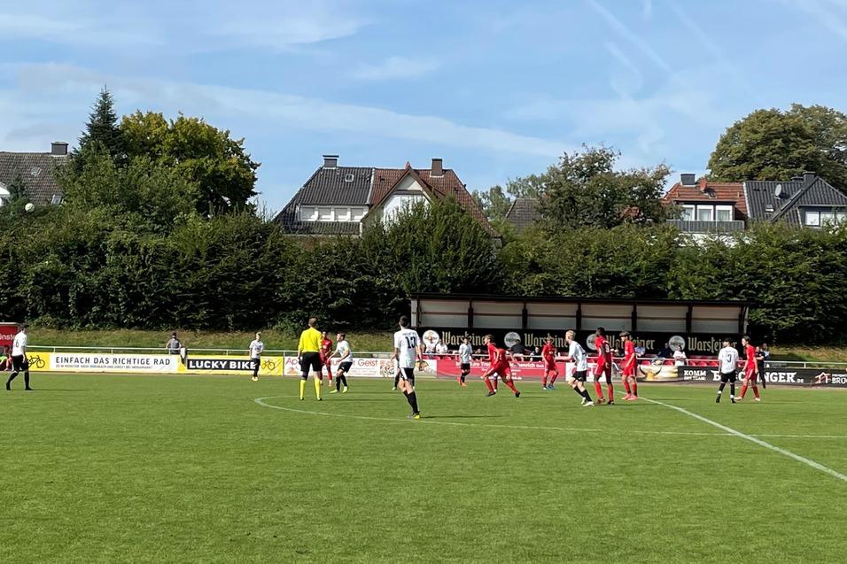 Fu&szlig;ballspiel auf gr&uuml;ner Wiese mit Spielern in roten und wei&szlig;en Trikots, im Hintergrund Wohnh&auml;user und B&auml;ume.