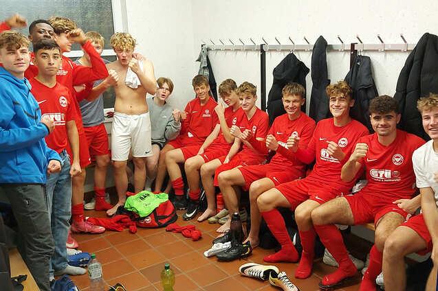 Jugendfu&szlig;ballmannschaft in roten Trikots sitzt in Umkleideraum, einige Spieler posieren und zeigen ihre Trikots.