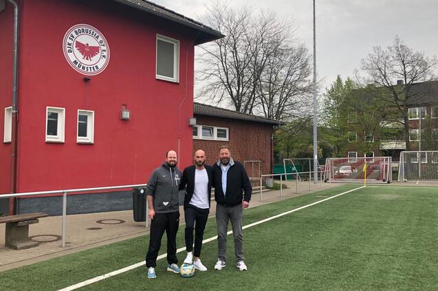 Drei M&auml;nner stehen auf einem Fu&szlig;ballplatz vor einem roten Vereinsgeb&auml;ude, mit einem Ball auf dem Rasen.