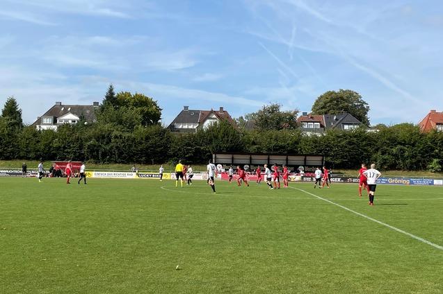 Fu&szlig;ballspiel auf gr&uuml;ner Wiese mit Spielern in roten und wei&szlig;en Trikots, im Hintergrund Wohnh&auml;user und B&auml;ume.