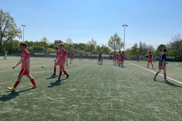 Fu&szlig;ballspiel mit Spielern in roten Trikots auf einem Kunstrasenplatz an einem sonnigen Tag.