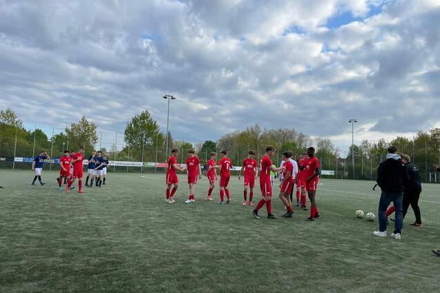 Fu&szlig;ballspieler in roten Trikots versammeln sich auf einem Kunstrasenplatz, im Hintergrund B&auml;ume und bew&ouml;lkter Himmel.