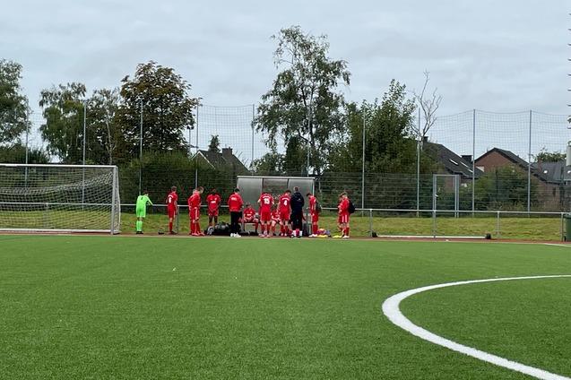 Eine Gruppe von Fu&szlig;ballspielern in roten Trikots versammelt sich an einer Auswechselbank auf einem Sportplatz.
