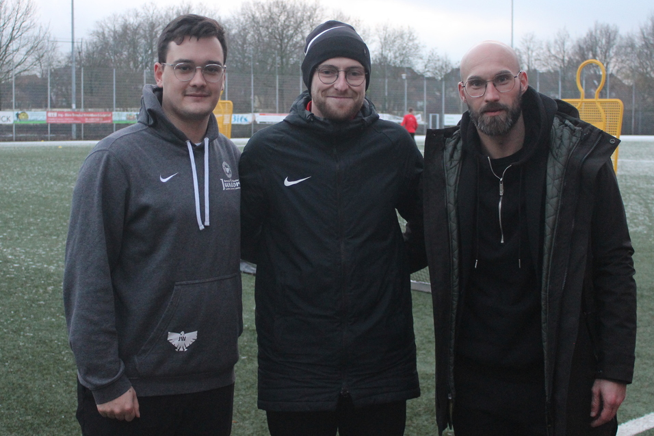 Drei Personen stehen auf einem gr&uuml;nen Fu&szlig;ballplatz, im Hintergrund sind Spieler und Fu&szlig;balltor sichtbar.