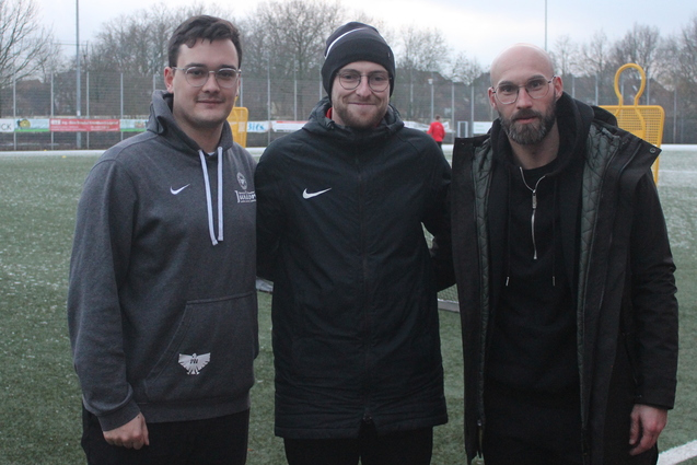 Drei Personen stehen auf einem gr&uuml;nen Fu&szlig;ballplatz, im Hintergrund sind Spieler und Fu&szlig;balltor sichtbar.