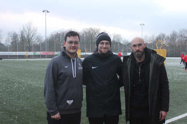 Drei Personen stehen auf einem gr&uuml;nen Fu&szlig;ballplatz, im Hintergrund sind Spieler und Fu&szlig;balltor sichtbar.