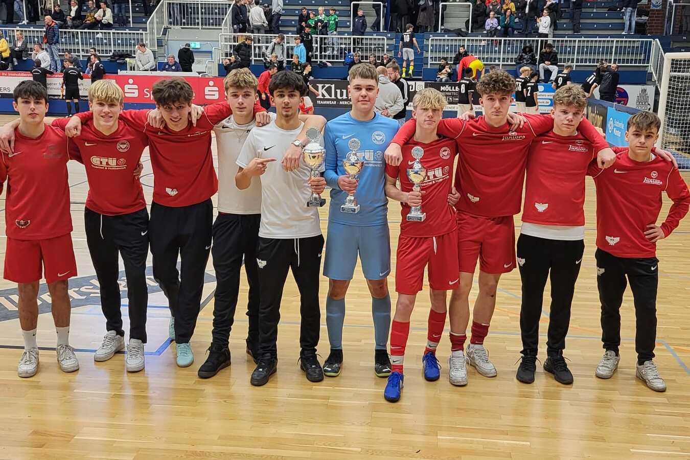 Gruppe von zehn jungen Fu&szlig;ballspielern in roten Trikots und Torwart in Blau posieren nach einem Spiel in einer Halle.
